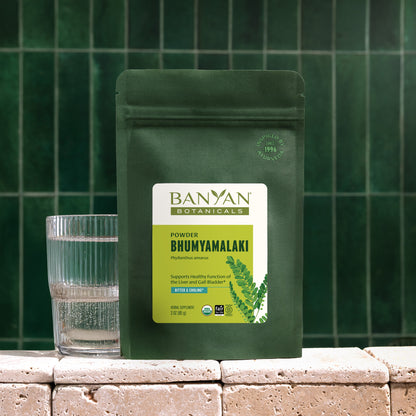 3 oz: Banyan Botanicals Bhumyamalaki powder pouch with a glass of water on a stone surface against a green tiled wall.
