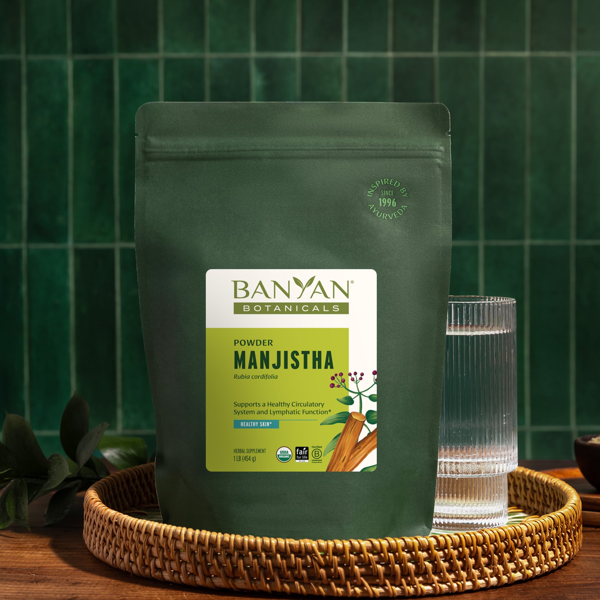 1 lb: Banyan Botanicals Manjistha powder package with a glass of water on a wooden surface.