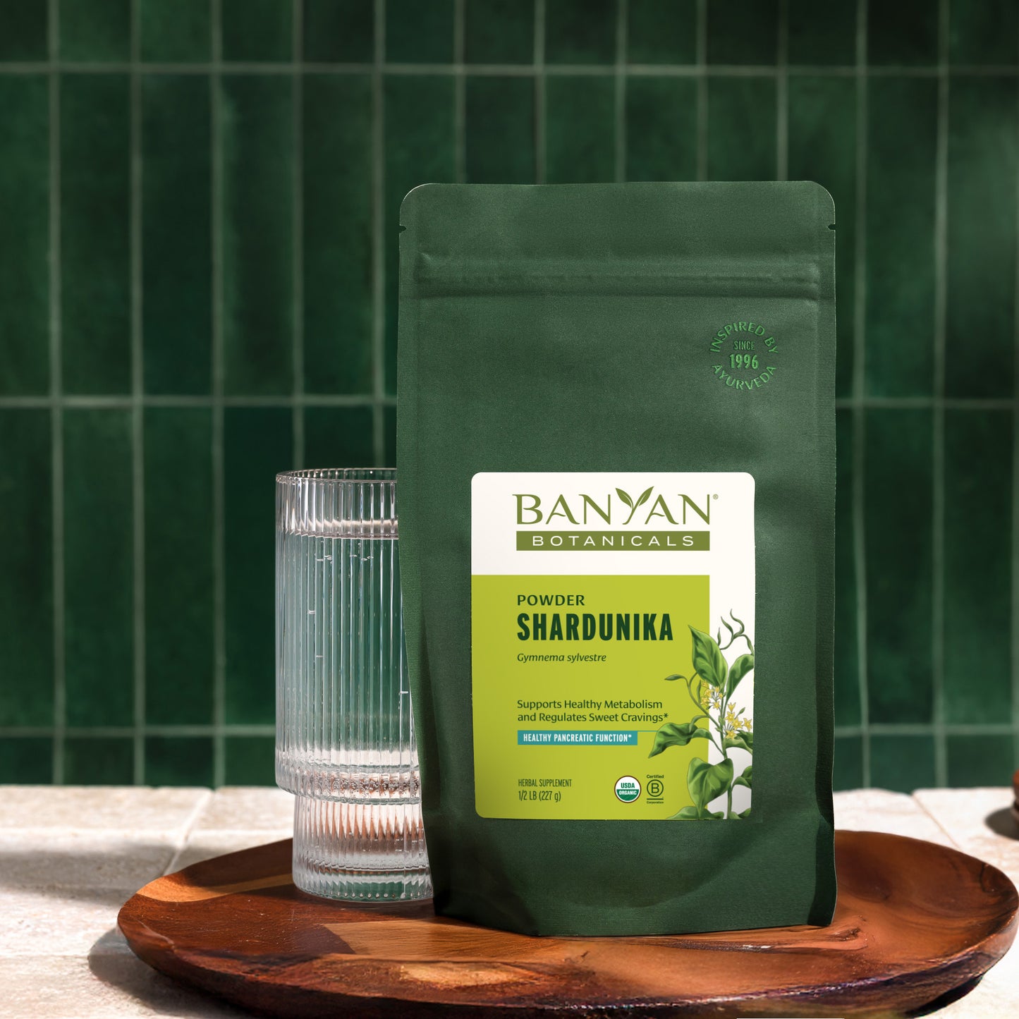 Banyan Botanicals Shardunika powder pouch with a glass on a wooden tray against a green tiled wall.